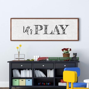 May include: A framed canvas print with the text "Let's Play" in black script font on a white background. The print is hanging on a white wall above a black bookshelf with toys and books.