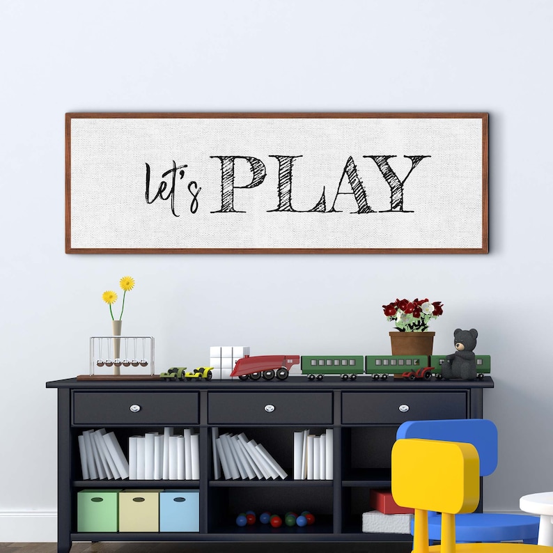 May include: A framed canvas print with the text "Let's Play" in black script font on a white background. The print is hanging on a white wall above a black bookshelf with toys and books.