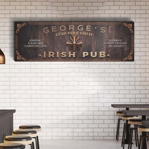 Puede incluir: Un letrero de madera con el texto "GEORGE'S C&eacute;ad Mile F&aacute;ilte IRISH PUB." El letrero tiene un tr&eacute;bol dorado con un borde dorado y el texto est&aacute; en una fuente dorada.