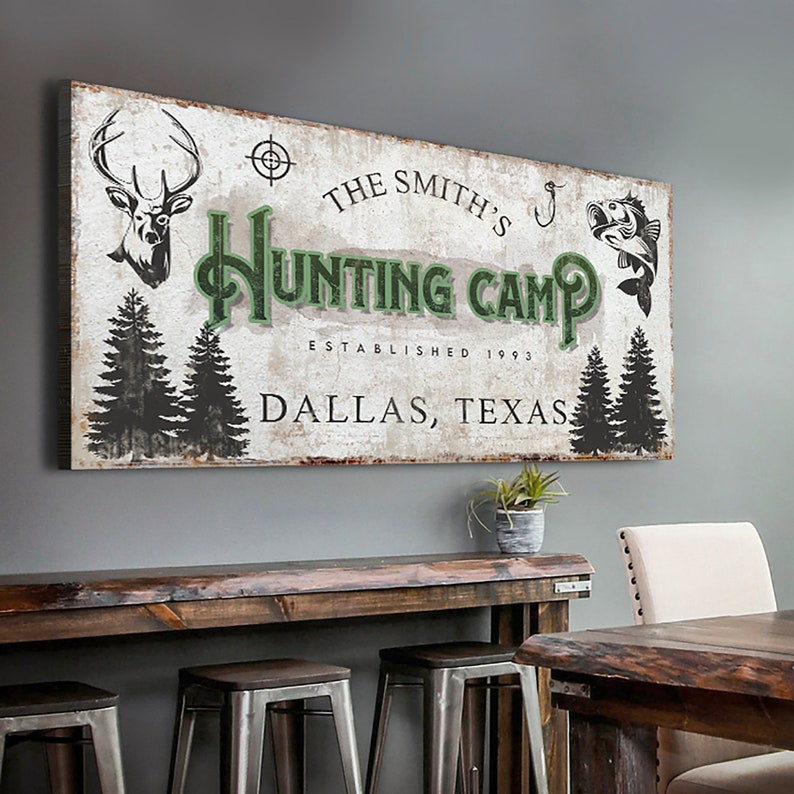 Puede incluir: Un letrero de madera r&uacute;stico con un fondo blanco desgastado y texto verde que dice "The Smith's Hunting Camp Established 1993 Dallas, Texas". El letrero presenta una cabeza de ciervo, un anzuelo de pesca y un pez.
