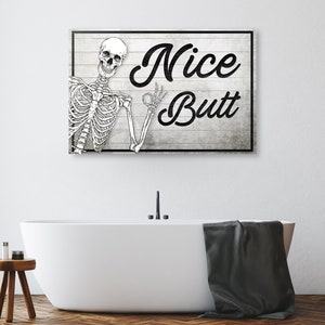 May include: A rectangular wooden sign with a black frame and the words "Nice Butt" in a bold, black script. A skeleton illustration is on the left, giving an "okay" hand gesture. The sign is mounted above a white bathtub.