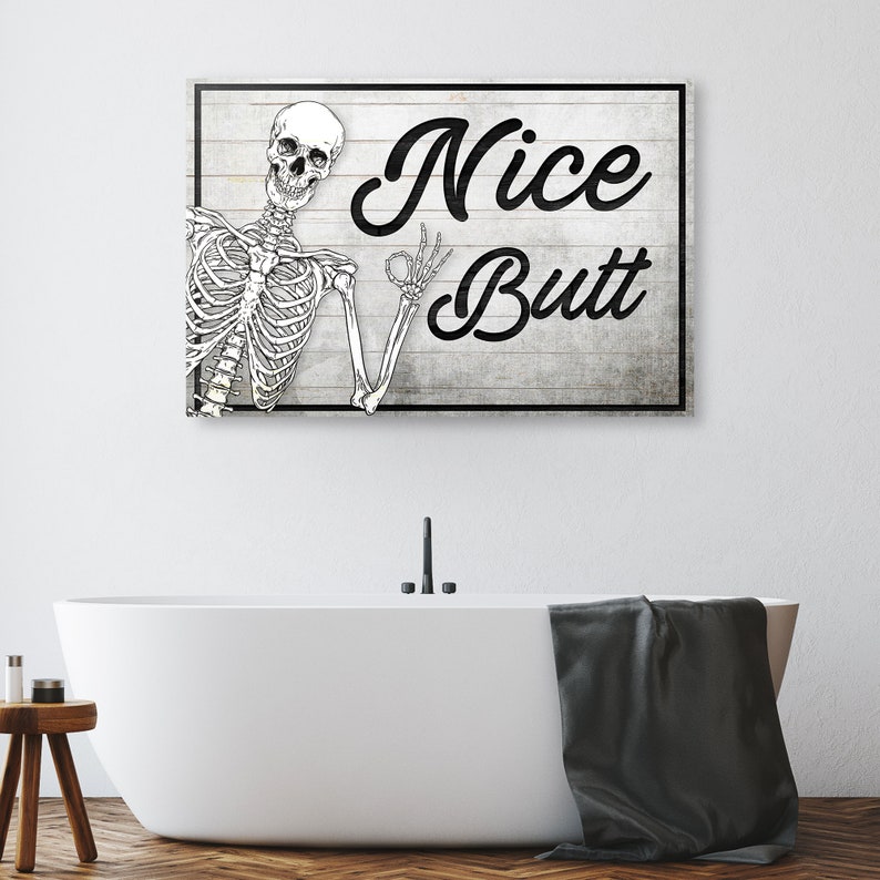 May include: A rectangular wooden sign with a black frame and the words "Nice Butt" in a bold, black script. A skeleton illustration is on the left, giving an "okay" hand gesture. The sign is mounted above a white bathtub.