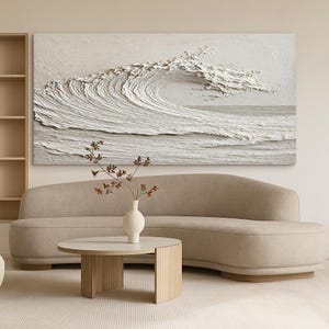 May include: A large, textured, white wave art piece hangs above a beige curved sofa. A round, light wood coffee table and a white vase with dried branches complete the living room scene. The artwork depicts a breaking wave.