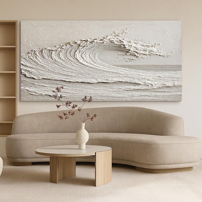 May include: A large, textured, white wave art piece hangs above a beige curved sofa. A round, light wood coffee table and a white vase with dried branches complete the living room scene. The artwork depicts a breaking wave.