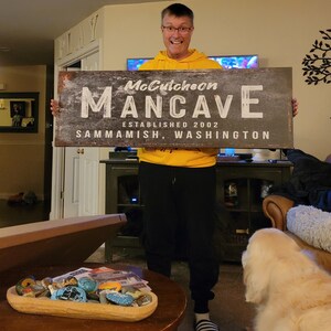 May include: A person holding a black and white sign that says "McCutcheon Mancave Established 2002 Sammamish, Washington".