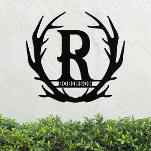 May include: Black metal monogram featuring a large letter "R" encircled by a ring of antlers. The name "ROBINSON" is inscribed in a smaller font beneath the letter.