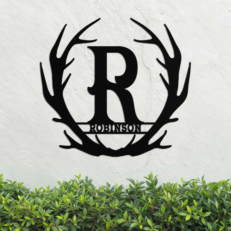 May include: Black metal monogram featuring a large letter "R" encircled by a ring of antlers. The name "ROBINSON" is inscribed in a smaller font beneath the letter.