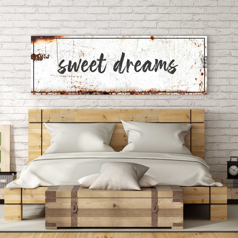 May include: A white metal sign with a rusty texture and black lettering that reads "sweet dreams". The sign is hanging on a white brick wall above a wooden bed frame.