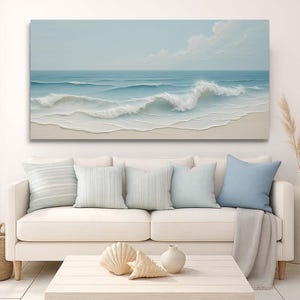 May include: A large rectangular canvas print depicting an ocean scene with waves crashing on a sandy beach. The artwork features shades of blue and white, with a light blue sky and fluffy clouds. The print hangs above a white sofa with light blue pillows.