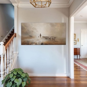 Puede incluir: Una pintura de paisaje rectangular representa a un pastor con un perro y un reba&ntilde;o de ovejas en una escena brumosa en tonos sepia. La obra de arte cuelga de una pared gris claro en un pasillo con una escalera y una planta de interior.
