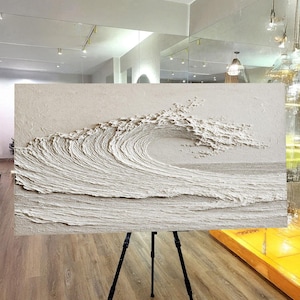 May include: A textured, off-white relief sculpture of a breaking ocean wave. The artwork is mounted on a black tripod, showcasing the wave's cresting form and the textured surface of the water. The piece is displayed in a well-lit room with a wooden floor.