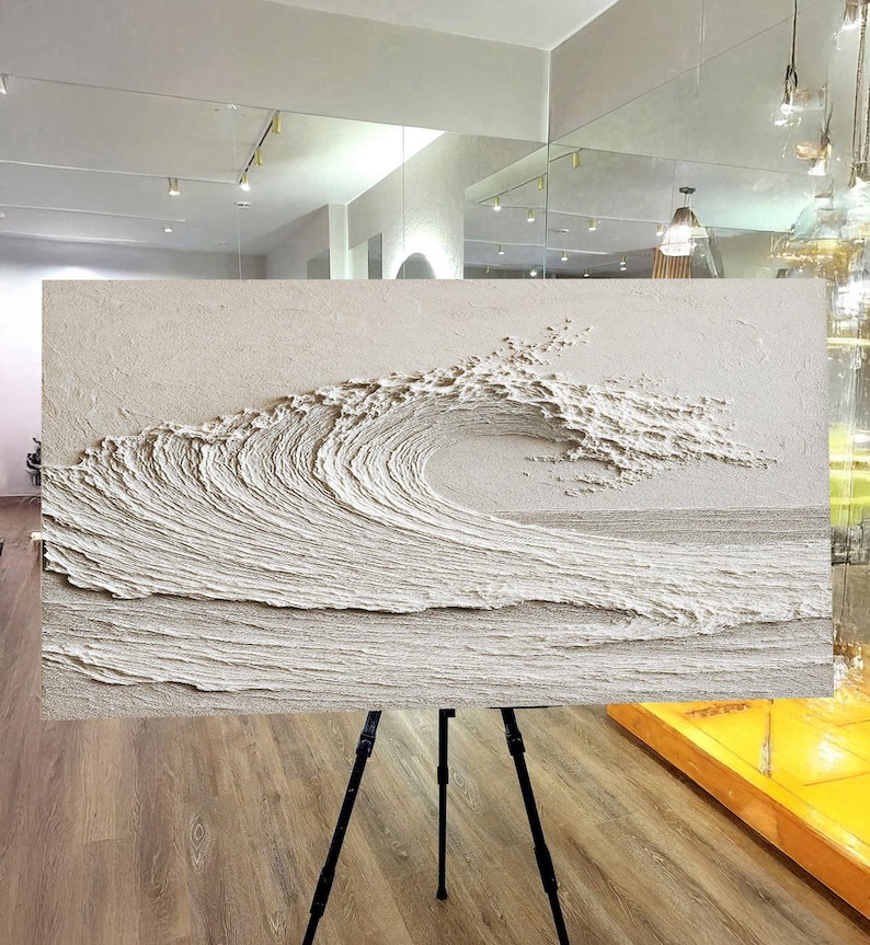 May include: A textured, off-white relief sculpture of a breaking ocean wave. The artwork is mounted on a black tripod, showcasing the wave's cresting form and the textured surface of the water. The piece is displayed in a well-lit room with a wooden floor.