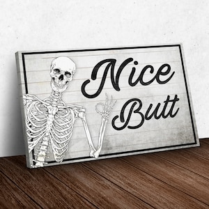 May include: A rectangular canvas print featuring a black and white skeleton illustration. The skeleton is giving an "okay" hand gesture. The words "Nice Butt" are written in a bold, black script on a gray wooden background.