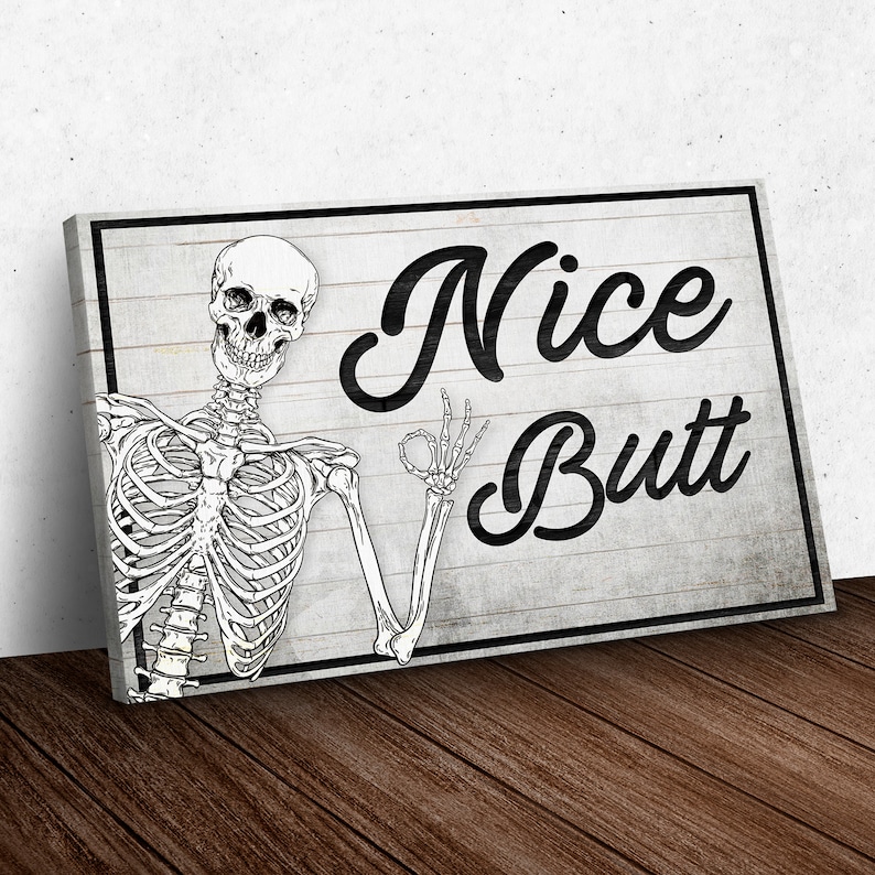 May include: A rectangular canvas print featuring a black and white skeleton illustration. The skeleton is giving an "okay" hand gesture. The words "Nice Butt" are written in a bold, black script on a gray wooden background.