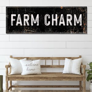 Puede incluir: Un letrero en blanco y negro con el texto "FARM CHARM" en un estilo vintage y desgastado. El letrero est&aacute; colgado en una pared blanca de tablones de madera sobre un banco de madera con almohadas.
