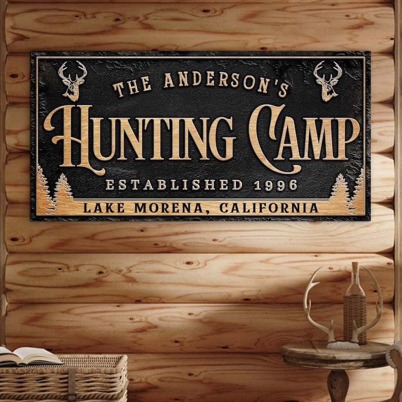 May include: Black and brown wooden sign with the text "The Anderson's Hunting Camp Established 1996 Lake Morena, California" with two deer head silhouettes.