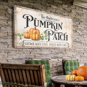 May include: A white sign with a rustic, vintage look, featuring the text "The Anderson's Pumpkin Patch" in black lettering with a decorative font. The sign also includes the text "Grown with love, picked with care" in smaller black lettering. The sign is decorated with a vine of green leaves and three orange pumpkins.
