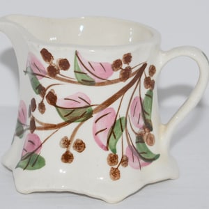 May include: A white ceramic creamer with a brown, green, and pink floral design. The creamer has a scalloped edge and a handle.