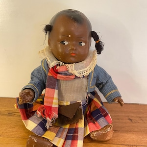 May include: A vintage doll with dark skin, painted facial features, and two small pigtails. The doll wears a blue jacket, a white lace-trimmed collar, a grey bodice, and a pleated skirt featuring a red, yellow, and blue plaid pattern.