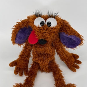 May include: A brown furry puppet with large white eyes, a red tongue sticking out, and purple ears. The puppet has long brown arms and legs.
