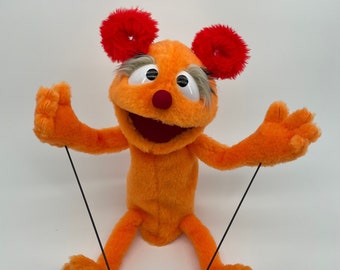 Puede incluir: Marioneta de mano naranja con orejas peludas rojas, cejas blancas y ojos grandes blancos. La marioneta tiene una nariz roja y una boca abierta de par en par. La marioneta está sentada con los brazos extendidos y está unida a dos cuerdas negras.