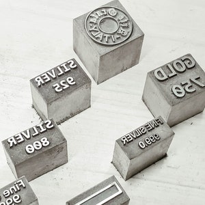 Può includere: Sei piccoli blocchi di cemento con diversi timbri metallici. I timbri indicano diverse finezze di oro e argento, tra cui 999 Oro fino, 999 Argento fino, 800 Argento, 925 Argento e 125 Oro.
