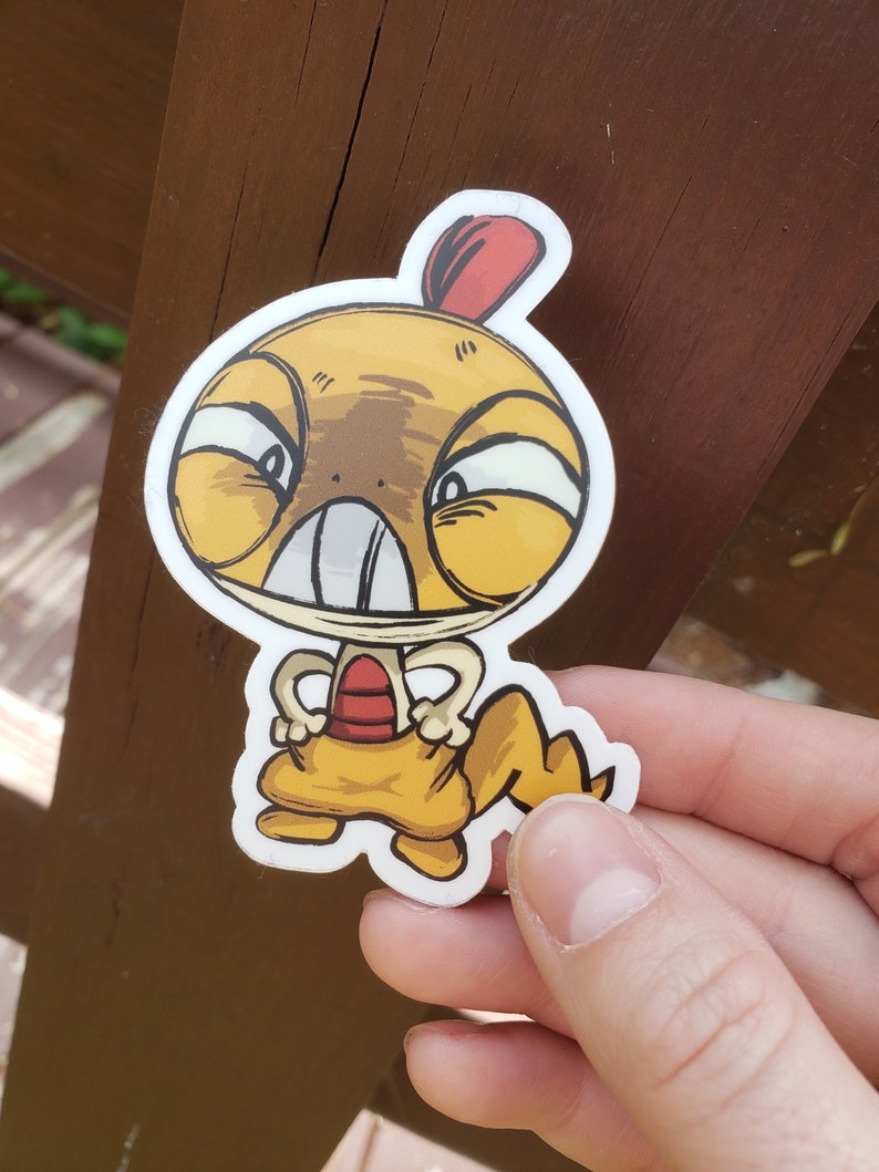 May include: A die-cut vinyl sticker featuring a cartoon character with a large yellow head, a red crest, and a wide mouth with prominent teeth. The character has a yellow body, a red chest patch, and a jagged tail, depicted in a bold graphic style.