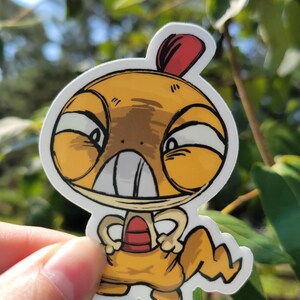 May include: A die-cut vinyl sticker featuring a cartoon character with a large yellow head, a red crest, and a tan body. The character has a wide, toothy grin, narrow eyes, and a jagged tail, rendered in a bold, black-outlined illustration style.