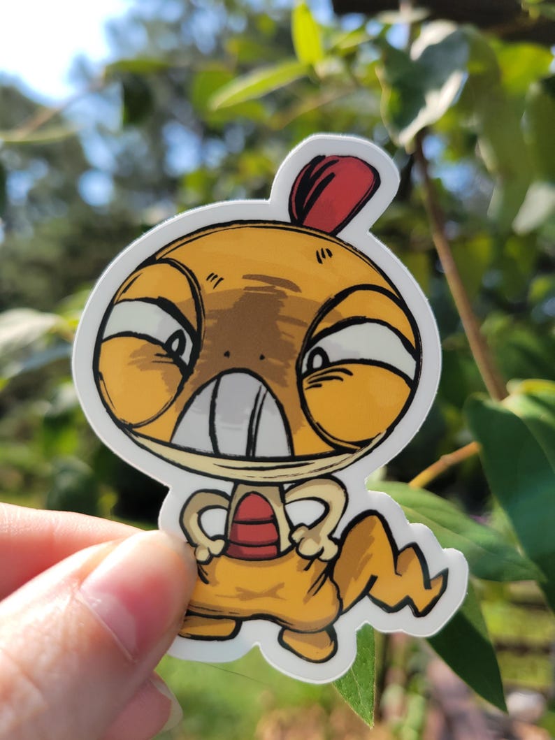 May include: A die-cut vinyl sticker featuring a cartoon character with a large yellow head, a red crest, and a tan body. The character has a wide, toothy grin, narrow eyes, and a jagged tail, rendered in a bold, black-outlined illustration style.