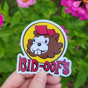 May include: A round sticker featuring a cartoon beaver wearing a red cap, set against a yellow circle. The sticker has the text "BID-OOF'S" in red and white. The sticker is held in a hand with a green background.