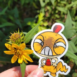 May include: A die-cut vinyl sticker featuring a cartoon character with a yellow, rounded head, large white eyes, a red crest, and a red chest patch. The character has a tan, lizard-like body with a jagged tail and a determined facial expression.