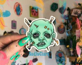 Puede incluir: Una pegatina con una cara de dibujos animados verde con pinceles que sobresalen de su cabeza. La cara tiene una expresión triste y dos estrellas en la frente. La pegatina está sobre un fondo blanco y tiene el texto "SadlyCiara" impreso debajo.