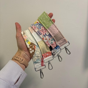 May include: A collection of colorful, patchwork fabric straps with silver-toned metal clasps. The straps feature various patterns, including floral, geometric, and plaid designs. Each strap is a unique combination of fabrics and colors.