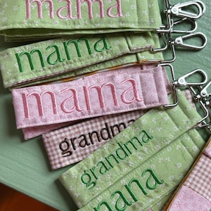 May include: Embroidered fabric keychains in various colors, featuring the words "mama", "grandma", and "nana". The keychains have silver-colored clasps and are made of patterned fabric in shades of green, pink, and brown.