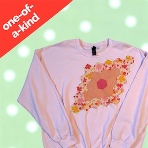 May include: A light purple sweatshirt with a floral patchwork design. The patchwork is a geometric shape with a pink center and is surrounded by yellow, pink, and red floral patterns. The text "one-of-a-kind" is in a red banner in the upper left corner.