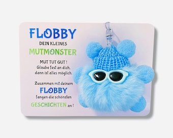Könnte beinhalten: Ein blauer, flauschiger Schlüsselanhänger mit einer Strickmütze und Sonnenbrille. Der Schlüsselanhänger trägt den Text "FLOBBY" und "MUTMONSTER". Der Text lautet außerdem "MUT TUT GUT! Glaube fest an dich, dann ist alles möglich. Zusammen mit deinem FLOBBY fangen die schönsten GESCHICHTEN an!"