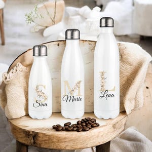 Könnte beinhalten: Drei weiße Edelstahl-Wasserflaschen mit Blumendesigns und personalisierten Namen: Sina, Marie und Lena.
