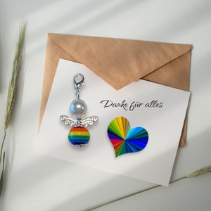 May include: A silver angel charm with a rainbow-coloured heart and a pearl bead. The charm is attached to a brown envelope with the text "Danke f&uuml;r alles" written on it.