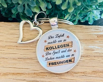 May include: Silver keychain with a heart charm and a round glass charm. The glass charm has a white background with brown text that reads "Der Zufall machte uns zu Kollegen. Der Spaß und das Lachen machte uns zu Freunden."