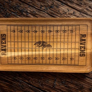 May include: A rectangular wooden serving tray featuring a laser-engraved American football field design. The tray displays yard line markings, the word RAVENS on both ends, and a central Baltimore Ravens team logo in the middle of the field.