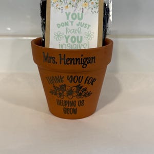 Puede incluir: Una maceta de terracota con texto negro que dice Mrs. Hennigan, THANK YOU FOR HELPING US GROW y un gráfico floral. Dentro de la maceta hay un paquete de plástico transparente con una tarjeta con flores amarillas y el texto YOU DON'T JUST TEACH YOU INSPIRE.