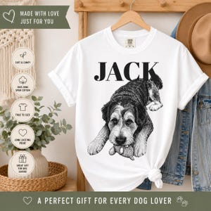 May include: A white short-sleeve t-shirt featuring a black and white graphic illustration of a dog lying down with a small toy in its paws. The name JACK is printed in large black capital letters above the dog. The shirt has a crew neckline.