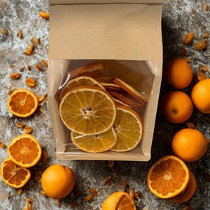 May include: A brown paper bag with a clear window, filled with dried orange slices. Fresh oranges and orange halves are scattered around the bag on a marble-patterned surface. The oranges are bright orange with a textured peel.