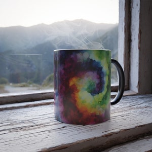 May include: A ceramic mug with a black handle, displaying a colourful watercolour design in shades of purple, blue, green, and yellow. Steam rises from the mug, suggesting a hot drink. The mug sits on a weathered, white-painted wooden windowsill.