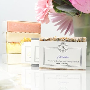 May include: A stack of handcrafted soaps in various colors, including pink, yellow, and gray, with a focus on a lavender-scented bar. The lavender soap is labeled "Unicorn Magic" and "Deluxe Handcrafted Soap - Mildly Scented", approximately 3.5 oz / 100 g.