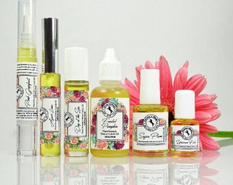 Puede incluir: Seis botellas de aceite esencial con aromas florales y frutales. Las botellas están etiquetadas con los nombres de las fragancias: Pomelo rosa, Lima Ayere, Día en el spa, Calabaza escarchada, Ciruela de azúcar y Polvo de unicornio. Las botellas están dispuestas en una fila sobre una superficie blanca con una flor rosa en el fondo.