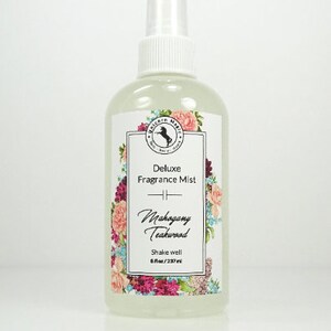 May include: A clear glass bottle with a white spray top and a white label. The label has a floral design and the text "Deluxe Fragrance Mist Mahogany Teakwood Shake well 8 fl oz/237 ml".