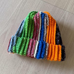 Può includere: Un berretto lavorato a maglia multicolore. Il berretto presenta strisce verticali in verde, blu, arancione, rosa e bianco. L'orlo è ripiegato e il berretto è realizzato in un filato morbido e strutturato.