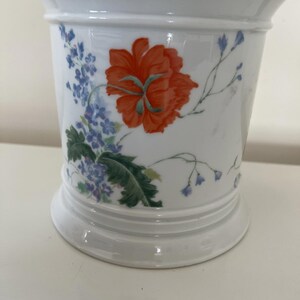 Könnte beinhalten: Weißer Keramik-Blumentopf mit ausgestelltem Rand und breitem Sockel. Der Topf ist mit einer großen orangefarbenen Blume und blauen floralen Akzenten verziert. Das Design umfasst grüne Blätter und zarte blaue Zweige.