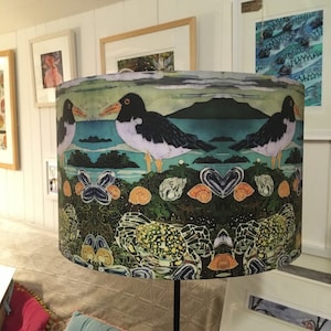 May include: A lampshade with a colourful design featuring two black and white birds, a crab, and seashells. The background is a watercolour-style landscape with a blue sky and green foliage.